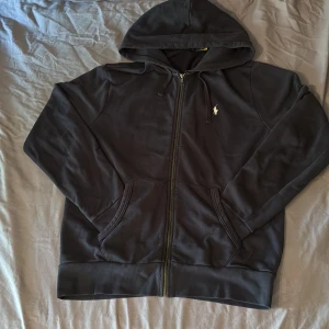 Svart/mörkblå hoodie från Polo Ralph Lauren - Svart hoodie från Polo Ralph Lauren med dragkedja och klassisk broderad logga på bröstet. Tröjan har huva med snörning och två fickor framtill. Tillverkad i mjuk bomull, perfekt för chill dagar eller när du vill ha en clean streetstyle.