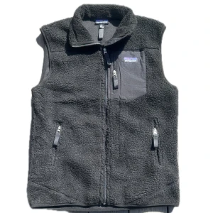 Svart fleeceväst från Patagonia - Mysig svart fleeceväst från Patagonia med hög krage och dragkedja framtill. Västen har tre fickor med dragkedja, varav en på bröstet med logga. Perfekt för lager-på-lager och chill stil.