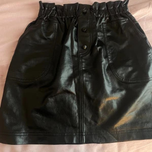 Svart skinnkjol från Funky Rocks - Cool svart kortkjol i skinnimitation från Funky Rocks. Kjolen har hög midja med resår och snygga knappar framtill samt stora fickor. Perfekt för dig som vill ha en edgy och trendig look.