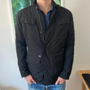 Svart parkajacka med dragkedja - Svart parkajacka med flera stora fickor framtill och hög krage. Jackan har dragkedja och tryckknappar, samt en stilren och enkel design. Perfekt för dig som vill ha en snygg och funktionell jacka till höst och vinter.