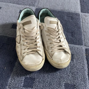 Vita Philippe Model sneakers i skinn - Snygga vita sneakers från Philippe Model med klassisk låg profil. Skorna har detaljer i vit och svart, snörning framtill och en rund tå. Tillverkade i skinn med ljusblått innerfoder och logotyp på sidan. Perfekta för en avslappnad och trendig look. Pris kan diskuteras!