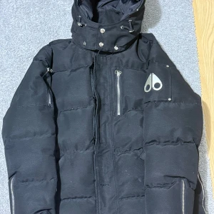 Svart pufferjacka Moose Knuckles M - Svart pufferjacka från Moose Knuckles i storlek M. Jackan har hög krage med snörning, flera dragkedjefickor och ikonisk metallogo på ärmen. Perfekt för kalla dagar med sin tjocka vaddering och stilrena look.  Observera: Dragkedjan är trasig, men kan enkelt fixas hos en skräddare för ca 100 kr.