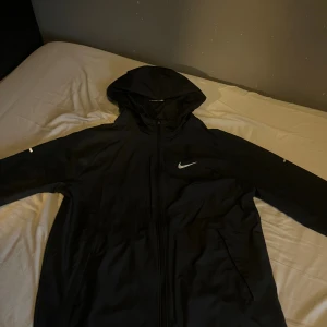 Svart Nike Miller vindjacka med huva - Svart vindjacka från Nike med reflekterande logga på bröstet och detaljer på ärmarna. Jackan har huva, hel dragkedja framtill och två sidofickor. Perfekt för löpning eller när du vill ha en sportig look. Tillverkad i lätt polyester.