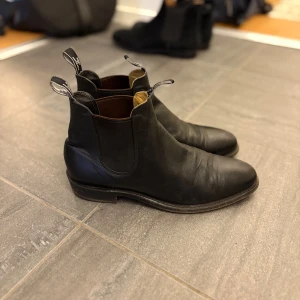 Svarta chelsea boots från R.M. Williams - Klassiska svarta chelsea boots från R.M. Williams i skinn med elastiska paneler på sidorna och dragflikar med logga. Snygg rund tå och låg klack, perfekt till jeans eller kostym. Skorna är stilren och tidlös design som funkar året runt. Priset kan diskuteras💸🪙