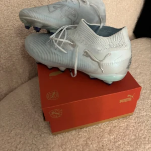 Puma Future 8 Pro Light Up fotbollsskor - Snygga ljusblå Puma Future 8 Pro Light Up FG/AG fotbollsskor med silvriga detaljer och stickad strumpliknande krage. Skorna har snörning och är tillverkade i syntetmaterial för bästa passform och grepp på planen. Använda en gång. 