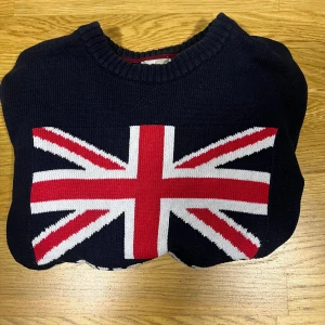 Mörkblå stickad tröja Newport - Mörkblå stickad tröja från Newport med Union Jack-flagga och texten 'NEWPORT' framtill. Tröjan har rund halsringning, ribbade muddar och är långärmad. Perfekt för dig som gillar klassisk brittisk stil och vill sticka ut.