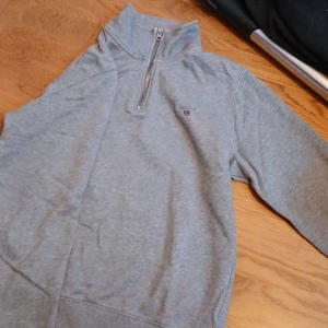 Grå half zip sweatshirt från GANT - Säljer en grå sweatshirt från GANT i storlek M. Tröjan har half zip-dragkedja, hög krage och broderad GANT-logga med sköld på bröstet. Tillverkad i mjuk bomull och har långa ärmar. Perfekt för lager-på-lager under kyliga dagar.
