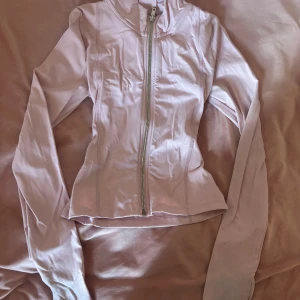 Ljusrosa träningströja med dragkedja - Snygg ljusrosa träningströja med hel guld dragkedja framtill och huva. Tröjan har långärmad design och är figursydd för en slim passform. Tillverkad i mjukt syntetmaterial som känns skönt mot huden. Perfekt för chill dagar eller när du vill ha en sportig look.