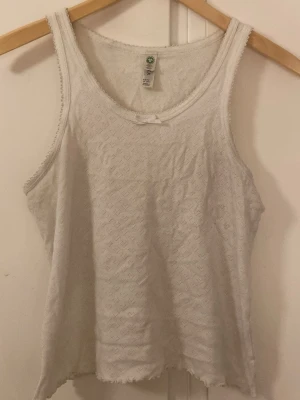 Vit spetslinne från Lindex - Sött vitt linnetopp från Lindex med spetskant och liten rosett framtill. Toppen har ett diskret hålmönster och är ärmlös med rund halsringning. Perfekt för varma dagar eller som lager under andra plagg.