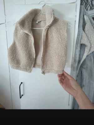 Beige teddyväst från NY Trend - Säljer en beige teddyväst från NY Trend i storlek S. Västen har en hög krage och dragkedja framtill. Den är croppad och gjord i mjukt fleece-material som ger en mysig vibe. Perfekt att slänga över en hoodie eller t-shirt för en chill look.