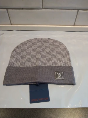 Grå rutig mössa från Louis Vuitton - Snygg grå mössa från Louis Vuitton med klassiskt rutigt mönster och LV-logga framtill. Mössan har en vikt kant och är tillverkad i mjukt material som passar perfekt för kyliga dagar.