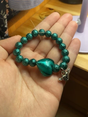 Grön pärlarmband med hjärthänge - Säljer ett coolt armband med gröna glittriga pärlor och ett stort hjärthänge i samma färg. Armbandet har även en liten silverfärgad berlock med vingar och nyckelhål. Perfekt accessoar för att lyfta din outfit.