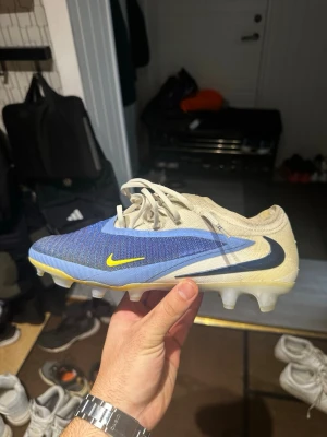 Nike Phantom fotbollsskor blå/beige - Nike Phantom fotbollsskor med stickad ovandel i blå och beige, detaljer i gult och svart. Skorna har snörning och platt sula med dobbar för gräsplan. Snygg design med rund tå och tydlig Nike-logga på sidan. Perfekt för dig som vill ha grym touch och kontroll på planen.
