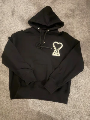 Svart hoodie från Ami Paris - Svart hoodie från Ami Paris med stor broderad hjärtdetalj och bokstaven A på bröstet. Tröjan har huva med snörning, magficka och ribbade muddar. Tillverkad i mjuk bomull, perfekt för chill dagar. Snygg och stilren design som sticker ut.