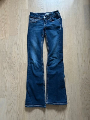 Blå bootcut jeans med broderad ficka -Gina 152 - Snygga blå jeans med bootcut-snitt och coola broderade detaljer på bakfickan. Jeansen har kontrastsömmar och klassisk femficksdesign. Materialet är jeans med en mjuk känsla och passformen är normal. Perfekt för dig som gillar lite extra detaljer på dina jeans.