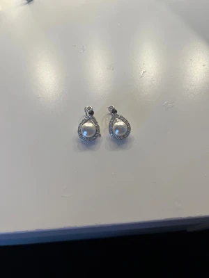 Örhänge  - Bilderna visar ett par pärlörhängen med droppformad design och små glittrande stenar, men ingen väst finns med på bilderna.