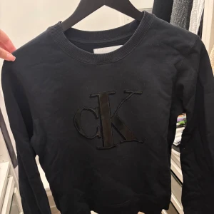 Svart sweatshirt från Calvin Klein - Säljer en svart sweatshirt från Calvin Klein med stor CK-logga framtill. Tröjan har rund halsringning och långa ärmar. Perfekt för dig som gillar stilrena och klassiska plagg med en twist.
