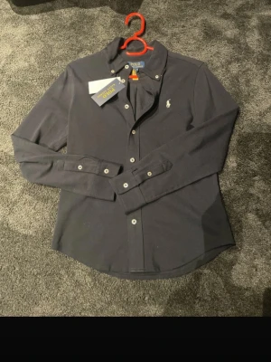 Mörkblå skjorta från Polo Ralph Lauren - Klassisk mörkblå skjorta från Polo Ralph Lauren med ikonisk broderad logga på bröstet. Skjortan har långa ärmar, knappar framtill och är tillverkad i mjuk bomull. Perfekt till jeans eller chinos för en clean och stilren look. Äkta ✅ , Ställ så mycket frågor ni vill 🌟