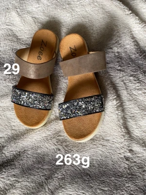 Glittriga sandaler från Zoee, strl 29 - Säljer ett par sandaler från Zoee i storlek 29. De har en bred rem i beige och en glittrig rem framtill med silverfärgade paljetter. Sulan är ljus och platt, och innersulan är mjuk och brun. Perfekta för sommarens alla äventyr!🌸Sparsamt använd. Stl 29