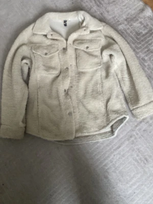 Beige teddyjacka med fickor - Mysig beige teddyjacka från H&M med två stora bröstfickor och knappar framtill. Jackan har en klassisk krage och långärmade ärmar, perfekt för att hålla värmen under kyliga dagar. Den har en loose passform och är tillverkad i mjukt fleece-material.