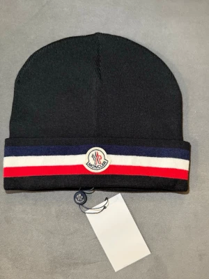 Svart Moncler mössa med ränder - Snygg svart mössa från Moncler med bred uppvikt kant och klassiska Moncler-loggan framtill. Mössan har en randig design i blått, vitt och rött längs kanten. Materialet är stickat och känns mjukt och bekvämt mot huvudet. Perfekt för dig som vill ha en stilren accessoar.