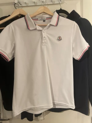 Vit Moncler piké med rödblå kant - Säljer en stilren vit pikétröja från Moncler med klassisk krage och korta ärmar. Pikén har Moncler-logga broderad på bröstet och snygga rödblå ränder på krage och ärmslut. Perfekt för dig som gillar sportig och clean look.