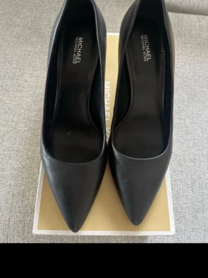 Svarta pumps från Michael Kors - Snygga svarta pumps från Michael Kors med spetsig tå och smal hög klack. Skorna är i skinn och har en klassisk, elegant form som passar till många olika outfits. Perfekta för dig som vill ha en stilren och tidlös look.