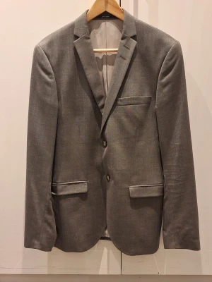 Grå kavaj från Tiger of Sweden - Tiger of Sweden kavaj i storlek 48 för herrar. Kavajen är grå och tillverkad av bomull. Den har en klassisk smal design med knappstängning och flera fickor fram. Superfint skick då den aldrig kommit till användning.  Pris 700 kr (nypris 4995 kr)