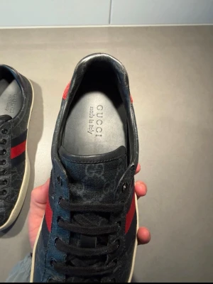 Gucci sneakers med röd och blå detalj - Säljer ett par snygga Gucci sneakers i svart med GG-mönster, röda och blå ränder på sidan samt röd, glansig häl med reptilmönster.