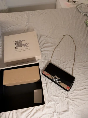Burberry bag - Säljer en snygg kuvertväska från Burberry med klassiskt rutig mönster i beige, svart, röd och vit. Väskan har en elegant guldfärgad kedja som axelrem och detaljer i svart skinn. Perfekt för dig som vill ha en ikonisk accessoar med lyxig känsla.
