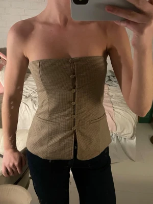 Beige rutig korsett med knappar - Säljer en beige axelbandslös korsett med rutigt mönster och dekorativa knappar framtill. Toppen har en liten slits nedtill och markerade sömmar som ger en snygg siluett. Perfekt att styla med jeans eller kjol för en trendig look.