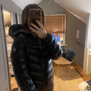 Svart pufferjacka från Colmar - Säljer en svart pufferjacka från Colmar med huva och dragkedja framtill. Jackan har en glansig yta och är vadderad i polyester, perfekt för kalla dagar. Klassisk design med långärmade ärmar och logotyp på armen. Snygg och enkel att matcha med olika outfits. Det är storlek 44 i Colmars storlekar vilket motsvarar ca XS/S