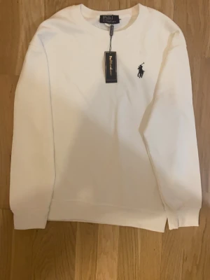 Vit sweatshirt från Polo Ralph Lauren - Vit sweatshirt från Polo Ralph Lauren i klassisk design med rund hals och svart broderad logga på bröstet. Tröjan har långa ärmar och ribbade muddar vid ärmslut och nederkant. Perfekt för en clean och stilren look.