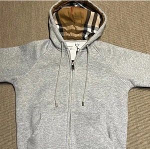 Burberry hoodie - Snygg grå hoodie från Burberry med klassiskt rutigt foder i huvan och broderad logga på bröstet. Tröjan har dragkedja, snörning i huvan och är tillverkad i mjuk bomull. Perfekt för dig som vill ha en stilren och lyxig look. 