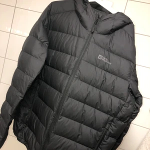 Svart dunjacka från Jack Wolfskin - Säljer en svart dunjacka från Jack Wolfskin med huva och dragkedja. Jackan är quiltad med breda kanaler och har två sidofickor med dragkedja. Perfekt för kalla dagar och riktigt skön att bära. Klassisk och stilren design.