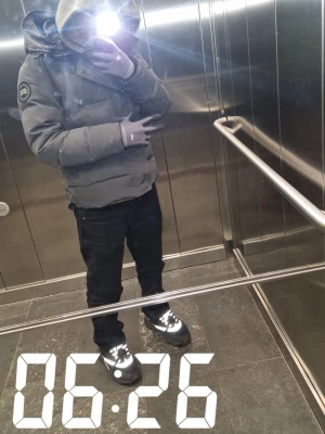 Grå pufferjacka från Canada Goose - Säljer en grå pufferjacka från Canada Goose med stor huva och logotyp på ärmen. Jackan har en clean look och är perfekt för kalla dagar. Den är långärmad och har en snygg, lite oversized passform. Grym att ha när det är kallt ute!