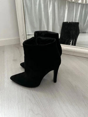 Svarta stövletter med klack från I Love Shoes - Säljer ett par svarta stövletter från I Love Shoes med spetsig tå och hög klack. Skorna är gjorda i mocka och har en stilren, elegant look som passar till jeans eller kjol. Perfekt för dig som gillar att sticka ut med snygga boots.