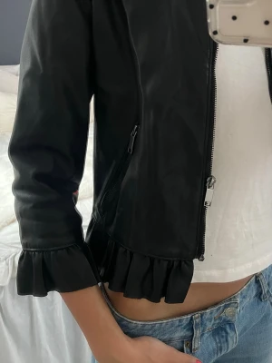 Svart skinnjacka med volang från Zara - Säljer en svart skinnjacka från Zara Basic med volangdetaljer vid ärmslut och nederkant. Jackan har rund hals, dragkedja framtill och är långärmad. Perfekt för dig som vill ha en edgy look med en twist. Fråga om du vill ha fler bilder eller info!