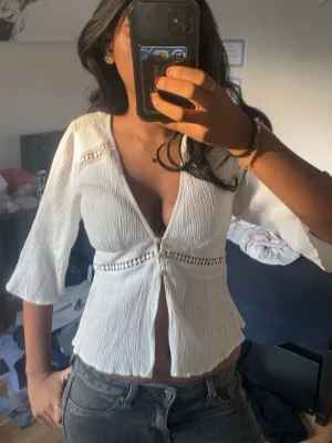 Vit blus med v-ringning och spetsdetaljer - Säljer en superfin vit blus med djup v-ringning och snygga spetsdetaljer vid axlar och midja. Blusen har trekvartsärmar och är tillverkad i ett lätt, krinklat tyg som ger en avslappnad känsla. Perfekt att styla med jeans eller kjol.