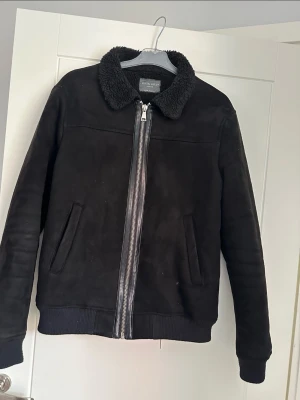 Svart teddyjacka från Primark stl M - Svart fodrad jacka mockaimitation från Primark med mjuk, fluffig krage och insida. Jackan har dragkedja framtill, två sidofickor och ribbade muddar vid ärmslut och nederkant. Perfekt för kalla dagar och har en stilren look som funkar till många outfits.  Stl M.  