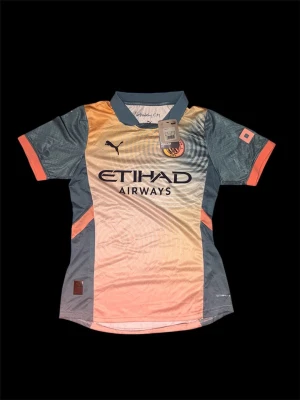 Manchester city tröja - Bilderna visar en Manchester City fotbollströja från Puma i färgerna blå, orange och beige med Etihad Airways-logga. Ingen sko finns på bilderna.