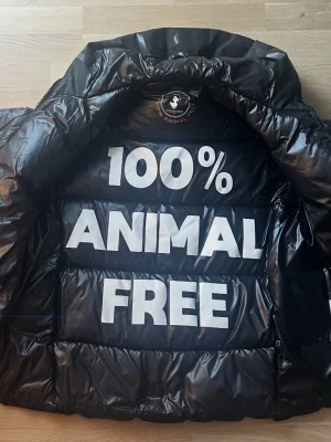 Svart pufferjacka ifrån Save The Duck - Säljer en svart pufferjacka från Save The Duck med huva och dragkedja. Jackan är quiltad och har två fickor med dragkedja. Insidan har ett stort tryck med texten '100% Animal Free'. Tillverkad i syntetmaterial och perfekt för kalla dagar. Storlek S men passar M.