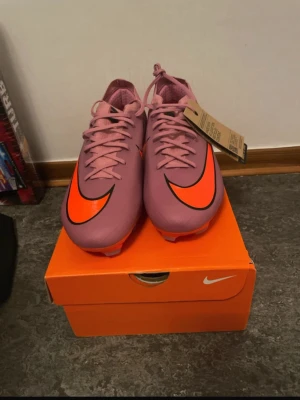 Nike Air Zoom fotbollsskor rosa/svarta - Säljer ett par Nike Air Zoom fotbollsskor i en snygg rosa färg med orangea och svarta detaljer. Skorna har snörning, platt sula och en rund tå. Materialet är syntet och skon har en stor M-logga på sidan. Perfekt för dig som vill sticka ut på planen.