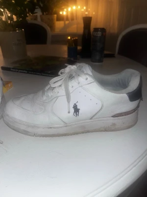 Vita sneakers från Polo Ralph Lauren - Säljer ett par vita sneakers från Polo Ralph Lauren med klassisk snörning och låg profil. Skorna har en rund tå, platt sula och detaljer i mörkblått på hälen där Polo-loggan syns tydligt. Materialet är skinn och insidan är mjukt vadderad för extra komfort.