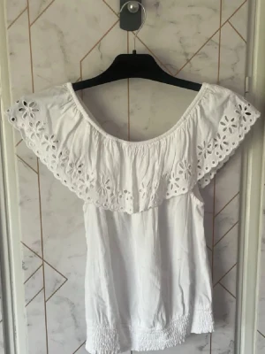 Vit offshoulder blus från H&M - Säljer en söt vit offshoulder blus från H&M med bred volangkant och broderade blommor. Toppen är ärmlös och har smockad nederkant för en bekväm passform. Perfekt till sommaren och ger en romantisk vibe. Superfint topp i storlek 170 men passar även xs och s.