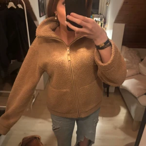 Beige/ brun teddyjacka med dragkedja - Mysig beige teddyjacka (kofta) med hög krage och hel dragkedja / zip up framtill. Jackan har två stora fickor och är tillverkad i ett fluffigt teddy-material som håller dig varm. Perfekt för dig som gillar en avslappnad och trendig stil.