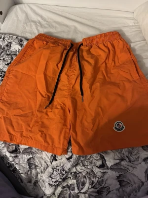 Orange Moncler shorts med snörning - Säljer ett par orangea shorts från Moncler med svart snörning i midjan och Moncler-logga på benet. Shortsen har en loose passform och är tillverkade i lätt syntetmaterial, perfekta för varma sommardagar. Fickor på sidorna och bak för extra funktion.