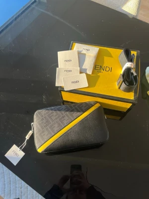 Fendi svart och gul axelväska - Säljer en snygg axelväska från Fendi i svart och grått med en diagonal gul detalj. Väskan har ett strukturerat mönster på ena sidan, justerbar axelrem och dragkedja upptill. Kommer med originalkartong och tags. Perfekt för dig som vill ha något unikt och trendigt.