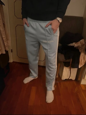 Ljusblåa Palm angels byxor - Dessa ljusblå Palm Angels track pants är både stilrena och bekväma. Med sina vita ränder längs sidorna ger de en sportig men ändå trendig look. Perfekta för en avslappnad dag eller för att piffa upp en enklare outfit! Ända defekten är en fett fläck som finns på bild 4 men inget som finns vid användning. Dessa byxor är även äkta! Skriv vid frågor eller funderingar😊
