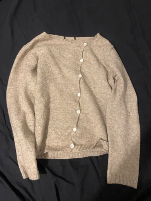 Beige kofta med sned knäppning - Säljer en beige kofta med lång ärm och sned knäppning med vita knappar framtill. Koftan är stickad och har ribbade muddar vid ärmslut och nederkant. Perfekt för dig som gillar en enkel men unik stil.